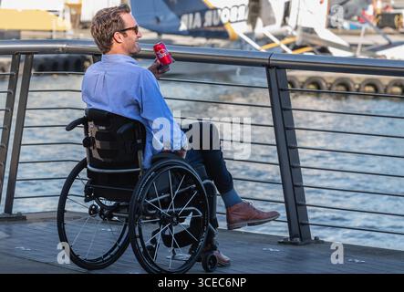 Disable man in wheelchair smiling and drinking coca cola. Young man in wheelchair dress in suit enjoying his life in urban city park. Inclusivity and Stock Photo