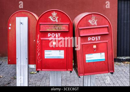 Three red post boxes, located outdoors on a paved urban street, against a red background wall. Stock Photo