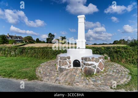 A white commemorative tar owenwith a stone foundation sits in a picturesque rural area, surrounded by fields, greenery, and a clear blue sky, evoking Stock Photo