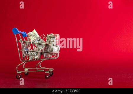 Shopping cart filled with shiny silver gift boxes, along with sparkling Christmas decorations, placed on a deep red background. Minimalist festive com Stock Photo