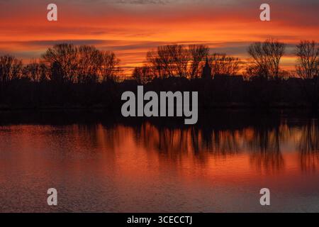 A stunning sunset over a calm lake with fiery red and orange skies reflecting in the water. The silhouettes of trees create a dramatic and peaceful la Stock Photo
