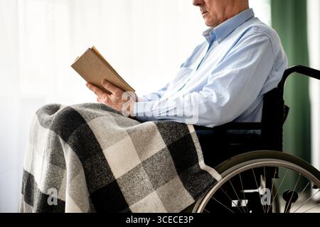 Cropped view of senior disabled man in wheelchair reading book indoors Stock Photo