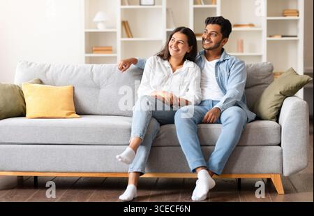 Happy dreamy indian couple looking aside at free space while sitting on sofa Stock Photo