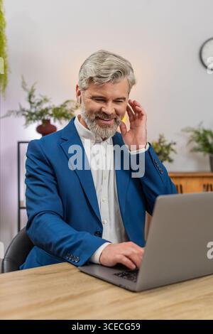Middle-aged businessman at home office codes on laptop wearing earphones listening to music while focusing on project. Freelancer guy at table enjoys it rhythm typing code delivering software feature. Stock Photo