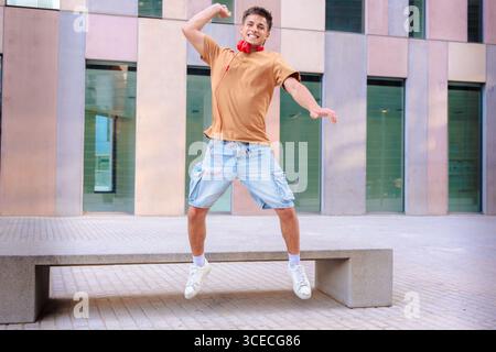 Happy young man jumping outdoors with arms raised in front of modern building Stock Photo