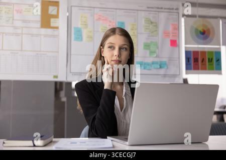 Thoughtful positive young project manager woman touching chin Stock Photo