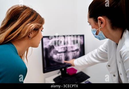 A dentist in a dental clinic shows an x-ray on a computer screen to a patient, explaining details while both focus on the image. The patient listens a Stock Photo