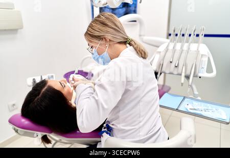 A dentist in a face mask conducts a dental check-up in a modern clinic. The patient is seated in a dental chair, surrounded by various dental instrume Stock Photo