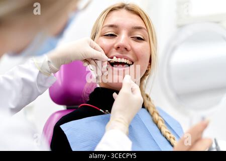 A dentist wearing gloves examines a smiling patient in a modern dental clinic. The patient holds a mirror while the dentist checks her teeth, highligh Stock Photo