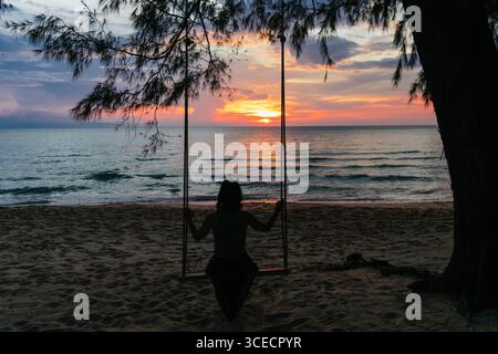 A woman sits on a swing overlooking the serene ocean at sunset on Koh Rong Island, Cambodia. The tranquil scene captures the vibrant sky and peaceful Stock Photo
