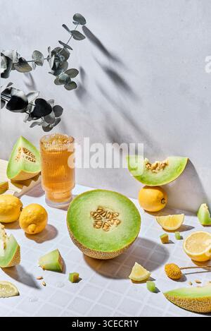 A vibrant display of fresh melons, lemons and fresh juice in a glass on a textured white background, highlighted by warm sunlight. The composition evo Stock Photo
