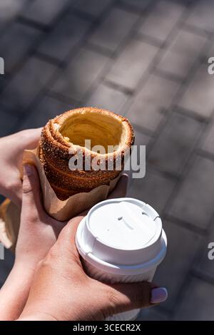 A boy holds a traditional trdelnik pastry in his hand, while a woman enjoys a cup of fresh coffee. The scene is set outdoors on a sunny day with a pav Stock Photo
