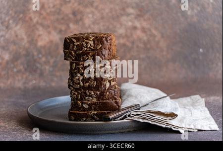Slices of nutritious whole wheat bread adorned with sunflower seeds, stacked neatly on a dark ceramic plate. A folded napkin and knife lie beside, con Stock Photo