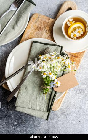 Top view of a summer dining scene with daisies on a napkin, silver cutlery, and a cup of tea with lemon. Set on wood and gray surfaces, it exudes a fr Stock Photo