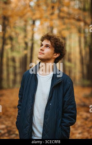 Young man standing in a Quebec forest during autumn, surrounded by colorful leaves. He wears a casual white T-shirt and blue jacket, looking up though Stock Photo