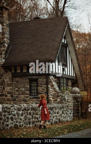 A woman in a red dress stands by a rustic stone house in Quebec, Canada, surrounded by autumnal trees. The cozy scene reflects the serene beauty of fa Stock Photo