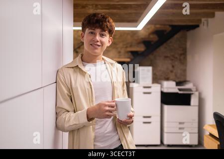 Young man enjoying a coffee break in a modern coworking space, standing relaxed with a cup. The office ambiance is inviting, fostering creativity and Stock Photo