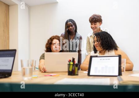 A diverse group of young coworkers collaborates around a laptop in a modern office setting. The environment is inclusive and professional, featuring t Stock Photo
