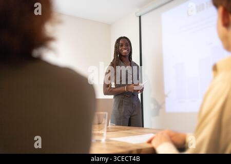 A, african woman is giving a presentation in a modern, multiracial office environment, engaging with colleagues seated around a conference table, focu Stock Photo
