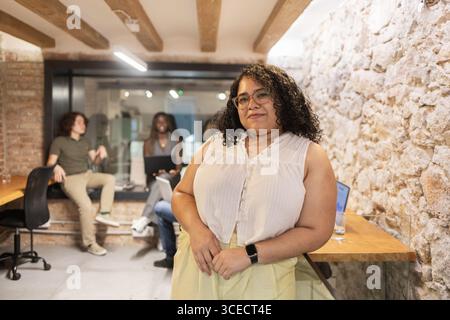 A confident female professional stands in the foreground of a modern office, while a diverse team collaborates in the background. Laptops and gadgets Stock Photo