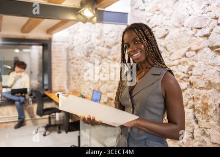 Young African woman with braids smiling while holding a document in a rustic coworking space. Another colleague works on a laptop in the background, c Stock Photo