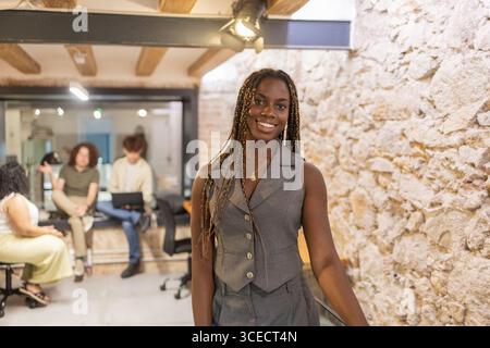 A young professional African woman with braids smiles confidently in a trendy summer coworking space, where colleagues engage in a lively discussion i Stock Photo