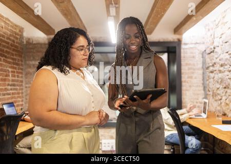 Two women are engaged in discussion, sharing ideas over a tablet in a modern coworking space. A person in the background is working on a laptop. Stock Photo