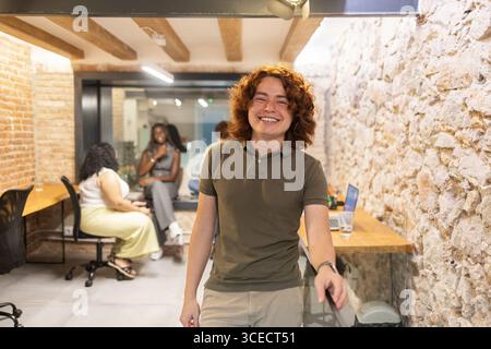 A young professional man smiles confidently in a lively coworking space with colleagues in the background having a discussion. A laptop on the table i Stock Photo