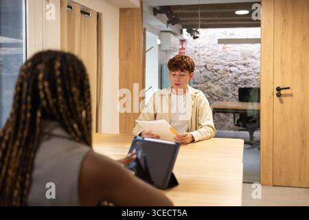 In a contemporary office, a diverse team collaborates. One person uses a digital tablet while another reviews documents, creating an inclusive workpla Stock Photo