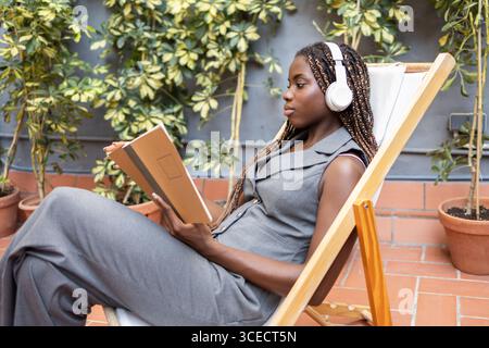 A young African woman with braids listens to music on headphones while relaxing with a notebook in a sunny coworking space, surrounded by lush plants Stock Photo