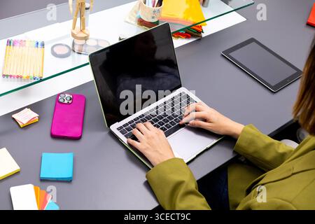 A professional workspace featuring a woman using a laptop at a clean and organized desk. The setting includes colorful stationery, a tablet, and perso Stock Photo