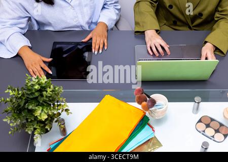 Two professionals collaborate using technology devices, surrounded by fabric swatches and makeup, during an image consulting session focused on color Stock Photo