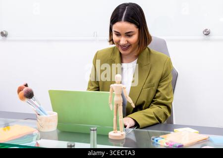 A professional image consultant works in a modern office setting, using a laptop. The desk is adorned with makeup brushes and a wooden mannequin, refl Stock Photo