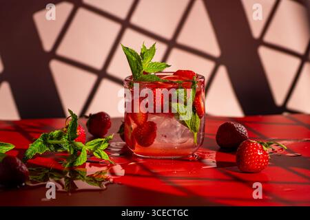 A vibrant strawberry mojito cocktail garnished with fresh mint leaves sits on a glossy red surface. The drink is surrounded by fresh strawberries, add Stock Photo