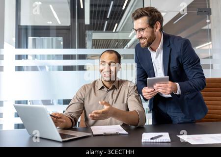 Two coworkers discuss ideas while working together using a laptop and tablet in an office. Stock Photo
