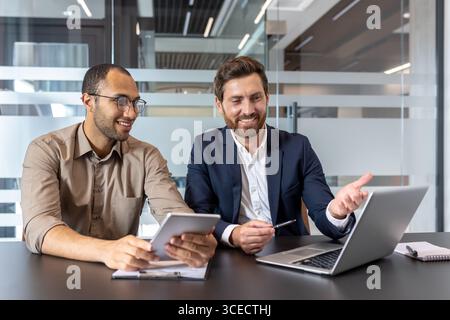 Business colleagues working together on a laptop and tablet, collaborating on a project in a well-lit, modern office environment. Stock Photo