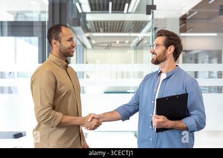 Two individuals engage in a handshake inside a contemporary office space, reflecting teamwork and partnership. The scene conveys a professional interaction and cooperation in modern environment. Stock Photo