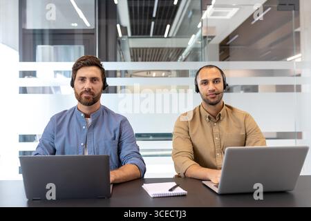 Two professionals wearing headsets work on laptops at a desk in office. The setting suggests customer service or technical support, reflecting collaboration, and a professional working environment. Stock Photo