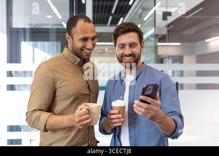 Two coworkers share a casual moment enjoying coffee while looking at a smartphone together in a contemporary office environment. This image captures teamwork, technology, friendly interactions Stock Photo
