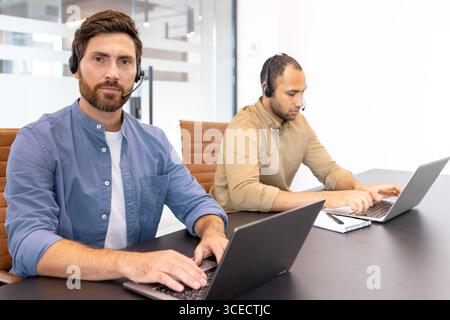 Two office professionals using laptops, wearing headsets, and engaged in a focused environment. They collaborate in an office setting, symbolizing communication, teamwork, and technology-driven Stock Photo