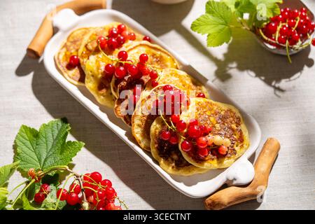 Top view of fluffy pancakes topped with vibrant red currants, elegantly arranged on a white ceramic plate. The light beige linen fabric adds a soft, r Stock Photo
