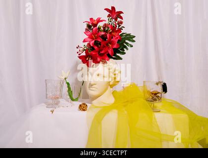 A contemporary still life scene featuring a classical bust adorned with vibrant pink flowers. Delicate glassware and a sheer yellow fabric drape the t Stock Photo