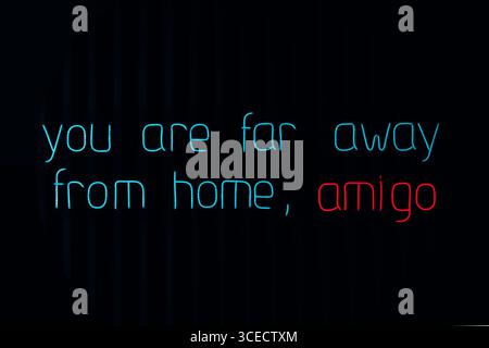 Neon sign with the phrase You are far away from home, amigo, in blue and red letters. The text is illuminated against a dark background, creating a st Stock Photo