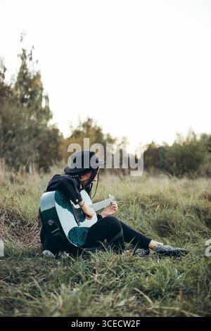 A woman sits on the grass, playing guitar, surrounded by the gentle glow of sunset. The scene captures a peaceful moment in nature. Stock Photo