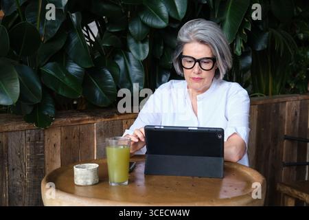 Mature woman with gray hair and glasses using a tablet at an outdoor cafe table with a refreshing green juice. She is focused on her digital work amid Stock Photo