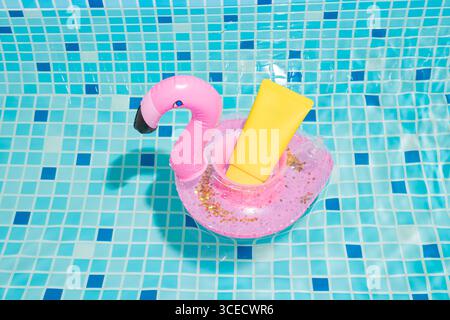 A yellow cosmetic sunscreen tube sits in a pink flamingo-shaped float in a sparkling pool. The vibrant aqua blue tiles create a refreshing summer back Stock Photo