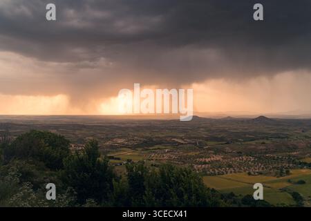 A dramatic storm unfolds over the Alcarria landscape at sunset, casting a warm glow over the vast fields. Dark clouds and distant rain create a striki Stock Photo