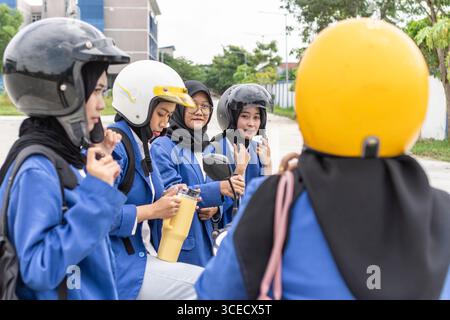 Group of Asian Muslim female students dressed in blue uniforms, wearing helmets, ready to travel to school on scooters. They appear engaged and prepar Stock Photo