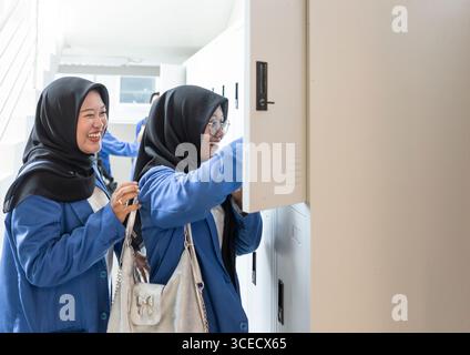 Two Asian Muslim students in school uniforms happily interact while organizing their belongings at lockers. Their cheerful expressions signify a posit Stock Photo