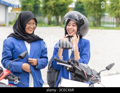 Two smiling Asian Muslim students in blue blazers stand beside their motorcycles, preparing for class with excitement as they start another school day Stock Photo